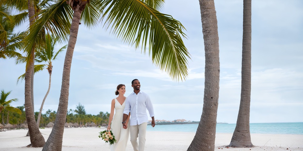 A couple dressed in wedding attire walks hand in hand along a tropical beach, highlighting the charm of budget-friendly destination weddings.