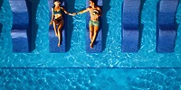 A couple celebrating their destination wedding relaxes on pool loungers at Hotel Riu Ventura, holding hands as they bask in the moment together, viewed from above.