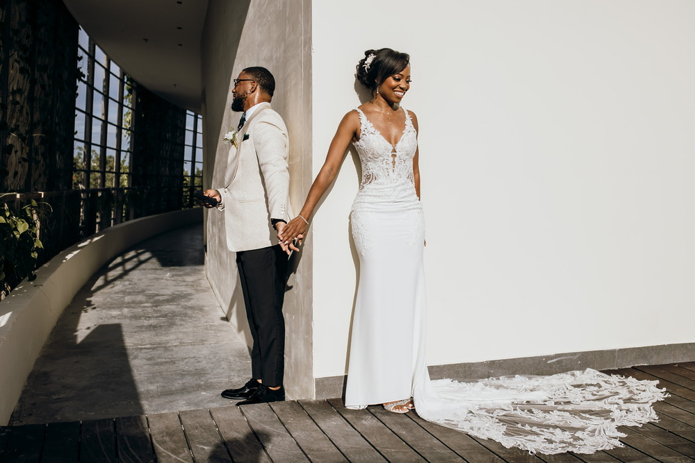 The bride and groom, celebrating their destination wedding, stand back-to-back around a corner, holding hands without seeing each other.
