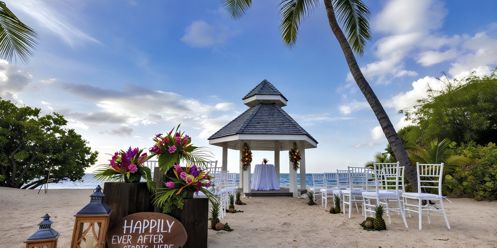 A beautiful destination wedding setup featuring a gazebo, white chairs, vibrant flowers, and a charming "Happily Ever After" sign arranged right on the beach.