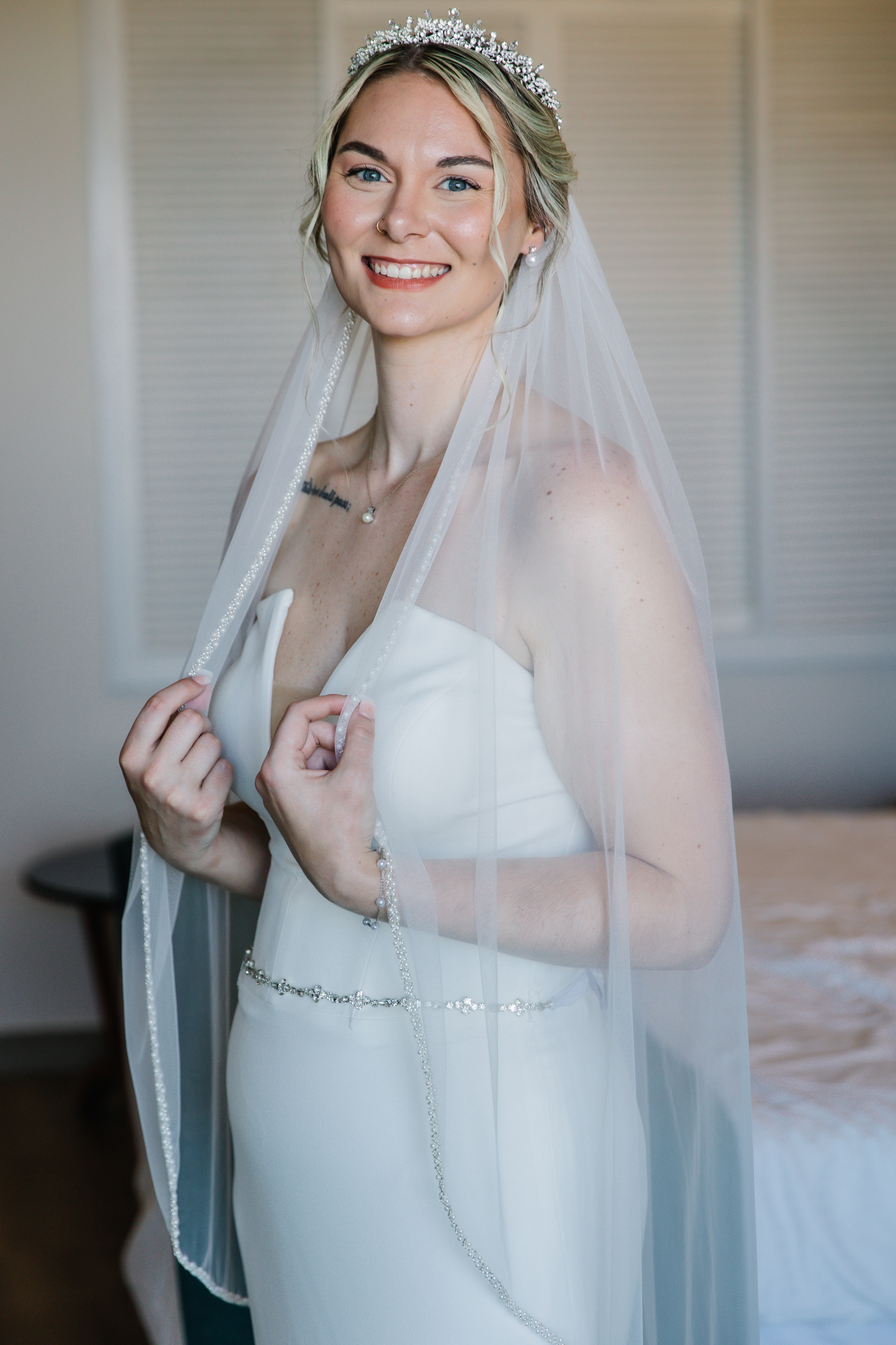 Carina and Steven: A bride in a white wedding dress and veil smiles indoors near a bed, capturing a joyful moment of their destination wedding.