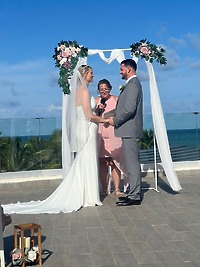 Carina and Steven hold hands beneath a floral arch at their oceanfront destination wedding.