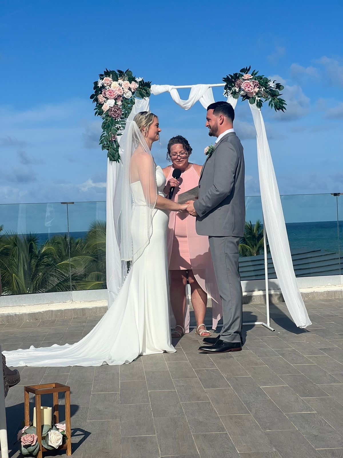 Carina and Steven hold hands beneath a floral arch at their oceanfront destination wedding.