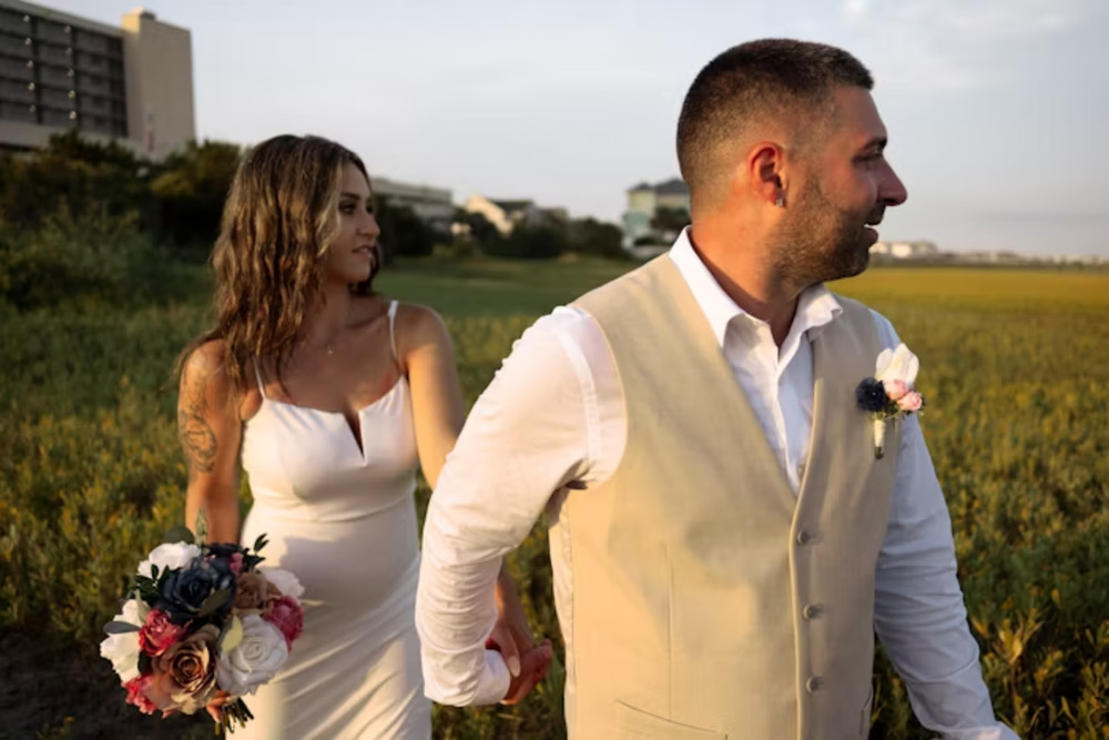 A bride and groom hold hands outside on a sunny day, standing together in a grassy area during their destination wedding.