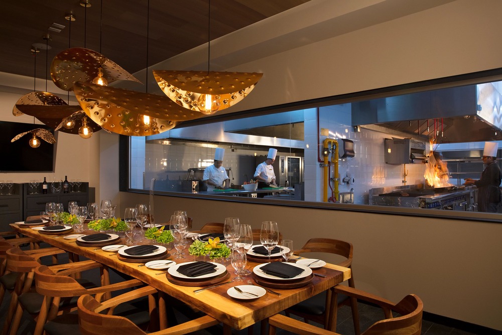 A modern dining room, elegantly set for a wedding celebration, overlooks a bustling kitchen through a large glass window—perfect for an intimate destination wedding dinner.