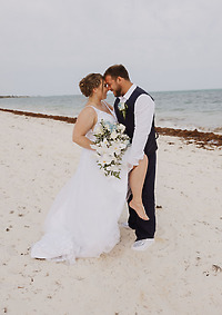 Ashleigh and Dominic embrace on the sandy shore, a bouquet in hand, with the ocean as their backdrop on their destination wedding day.