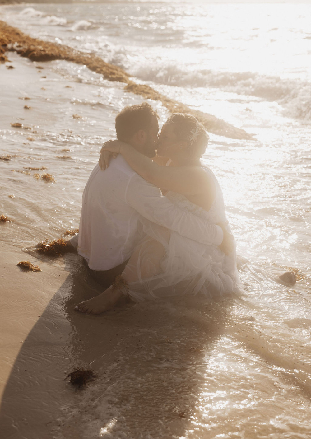 Ashleigh and Dominic, elegantly dressed for their destination wedding, embrace on the shoreline amidst gentle waves and scattered seaweed.