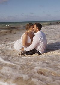 Ashleigh and Dominic share a kiss on the shore, surrounded by ocean waves, as their destination wedding unfolds in paradise.