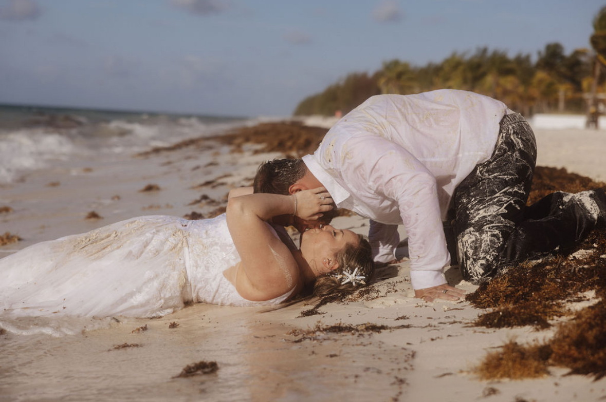 Ashleigh and Dominic, dressed in wedding attire, lie together on a seaweed-strewn beach by the ocean, capturing the essence of a destination wedding.