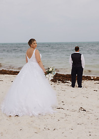 Ashleigh and Dominic: The bride, dressed in a stunning white gown and holding her bouquet, stands on the beach behind the groom at their destination wedding.