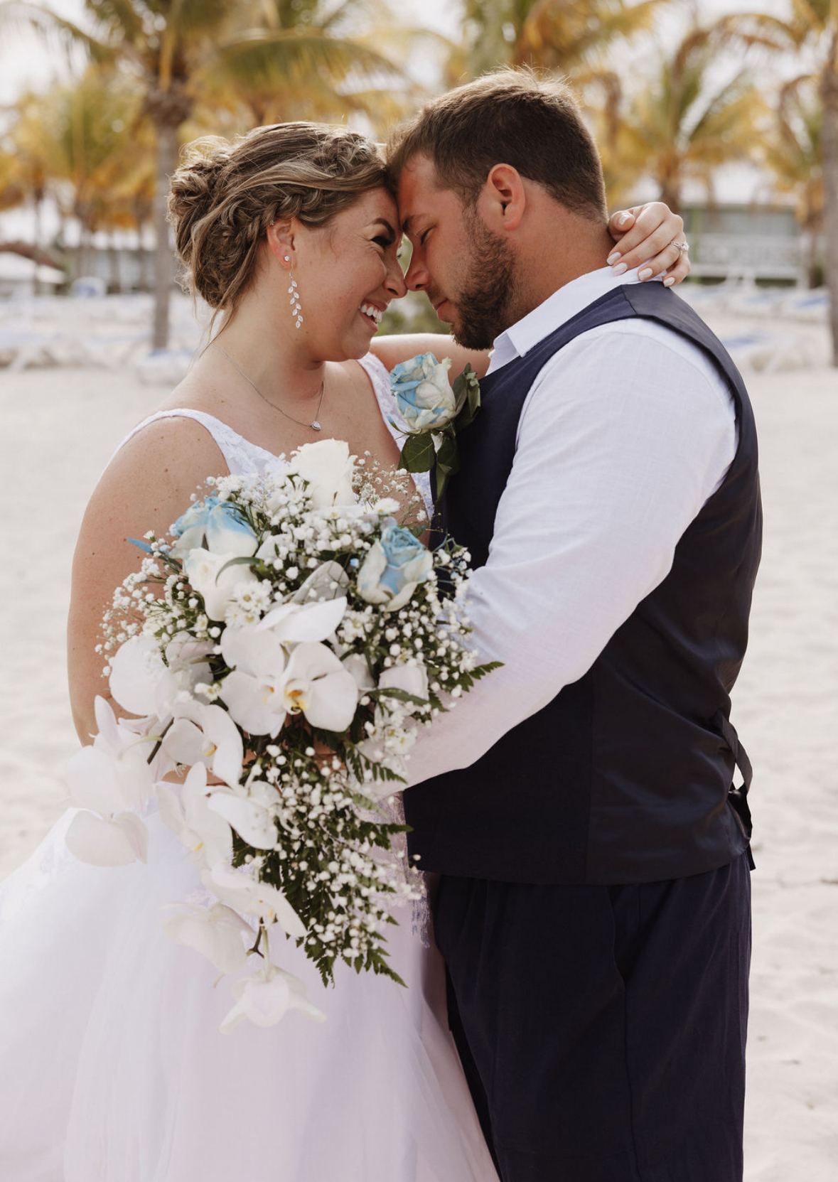 Ashleigh and Dominic share an embrace on the beach during their destination wedding, holding a bouquet of white and blue flowers.