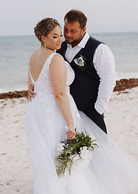 Ashleigh and Dominic stand together on the beach during their destination wedding, with the bride holding a bouquet of white flowers.