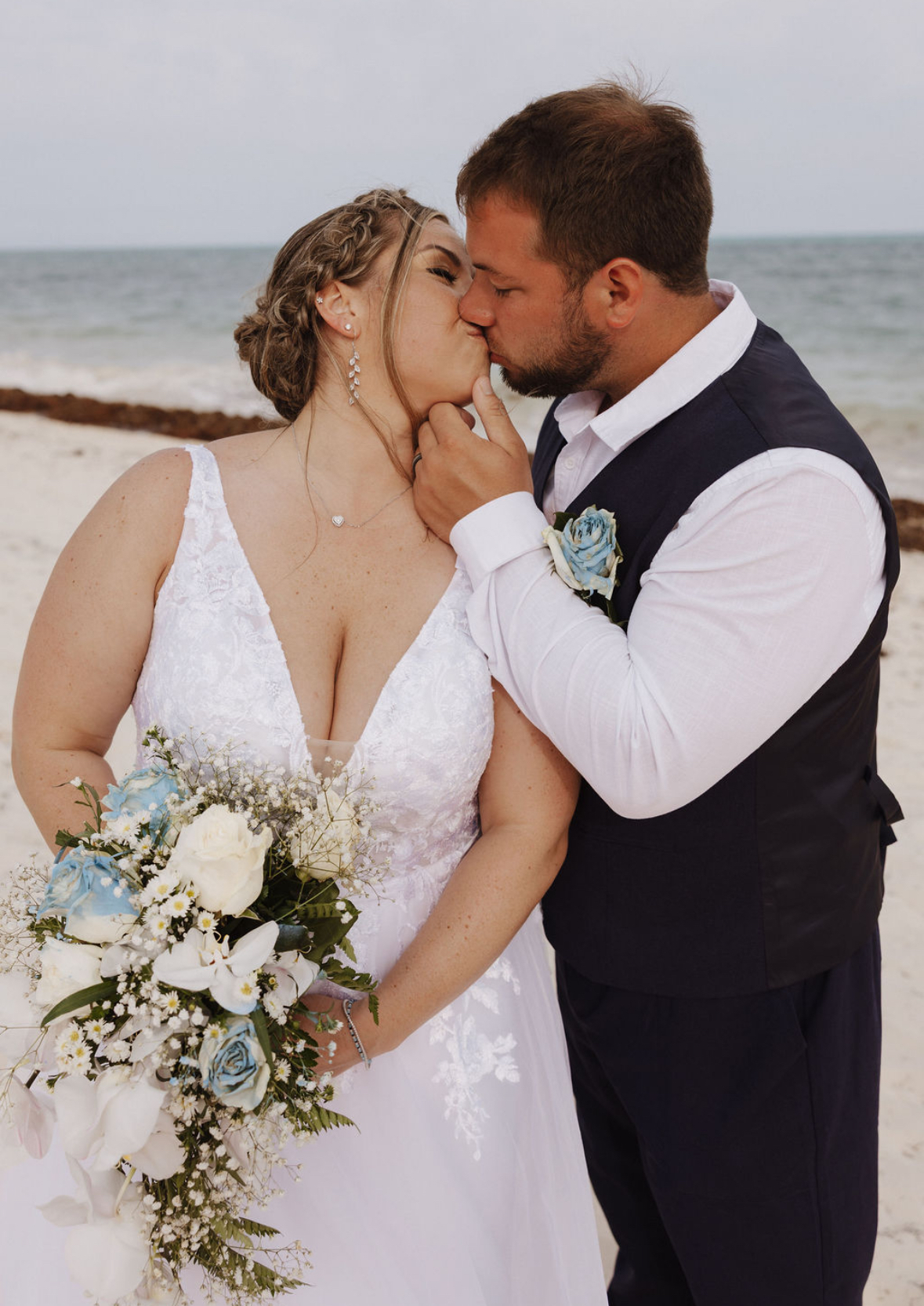 Ashleigh and Dominic share a romantic kiss on the beach during their destination wedding, with the bride holding a beautiful blue and white bouquet.