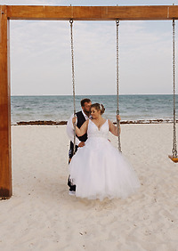 Ashleigh and Dominic pose together on a beach swing, with the ocean glistening in the background, capturing a beautiful moment from their destination wedding.