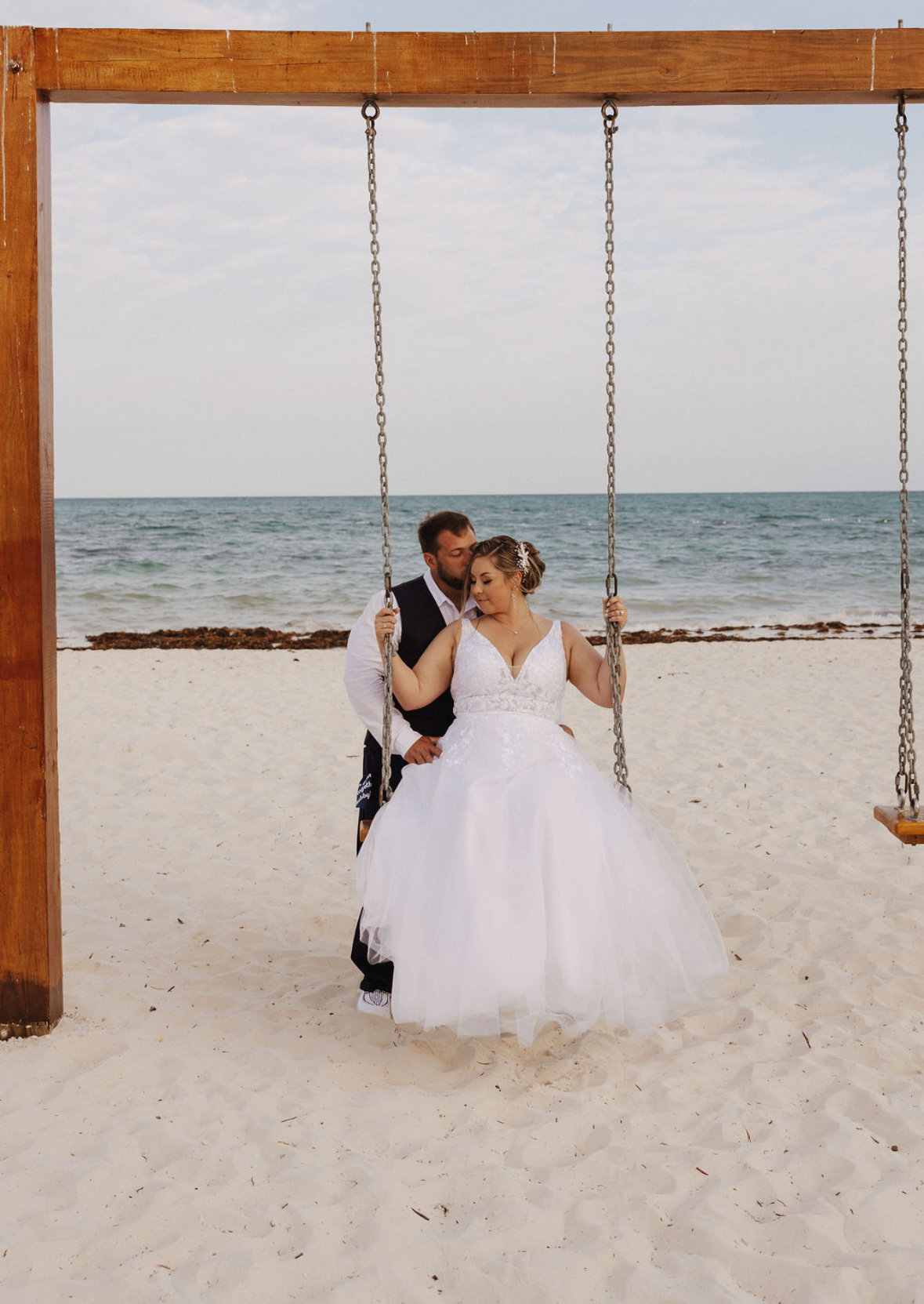 Ashleigh and Dominic pose together on a beach swing, with the ocean glistening in the background, capturing a beautiful moment from their destination wedding.