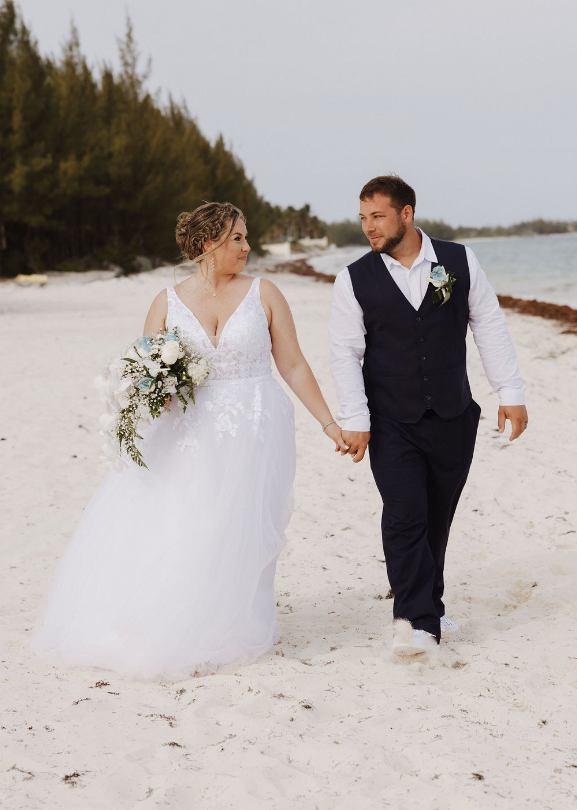 Ashleigh and Dominic, hand in hand, stroll along a sandy beach in their elegant wedding attire, the perfect scene for a destination wedding.