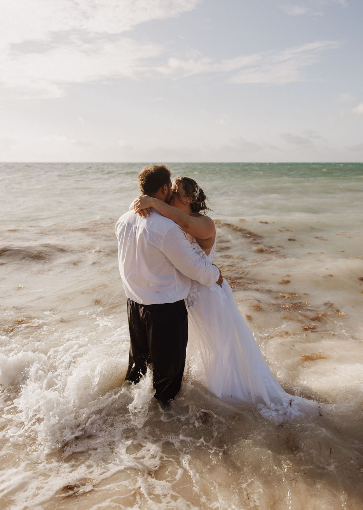 Ashleigh and Dominic share a kiss in their wedding attire, standing together in the shallow ocean waves during their destination wedding.