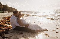 Ashleigh and Dominic, dressed in their wedding attire, sit together on a sandy beach near the water’s edge, surrounded by seaweed—capturing a beautiful moment from their destination wedding.