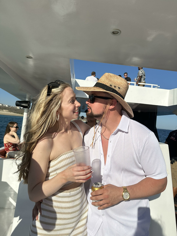 At a destination wedding, a man and woman stand close together on a boat, holding drinks, with other guests mingling in the background.