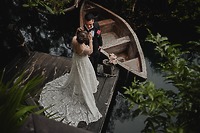 Nancy and David stand on a dock beside their rowboat, water shimmering behind them and lush greenery all around—the perfect backdrop for their destination wedding.