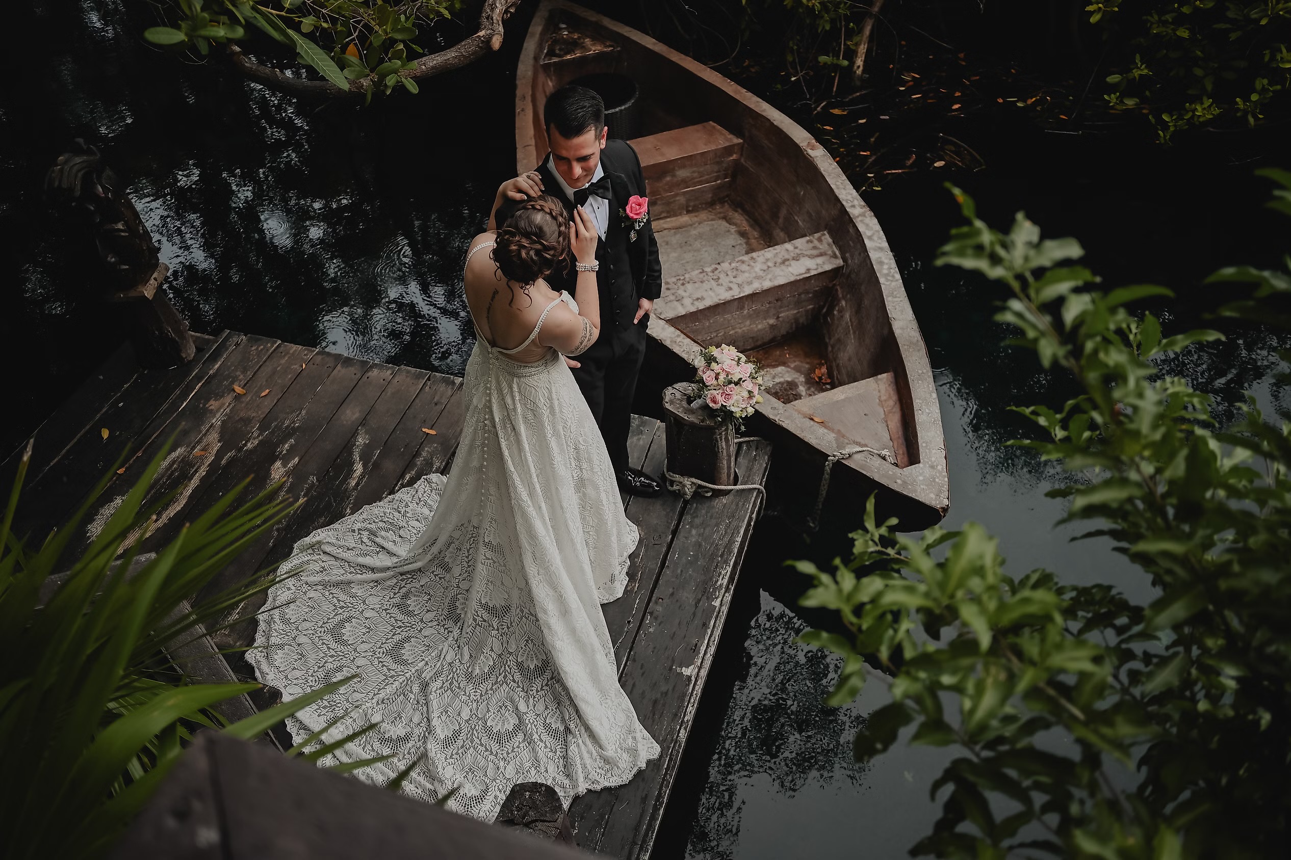 Nancy and David stand on a dock beside their rowboat, water shimmering behind them and lush greenery all around—the perfect backdrop for their destination wedding.