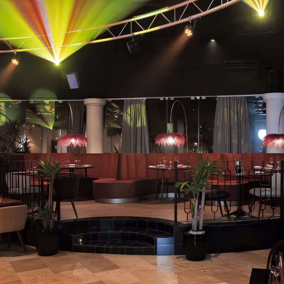 A modern restaurant interior, perfect for a destination wedding celebration, featuring vibrant stage lighting, elegant red booths, and lush potted plants.