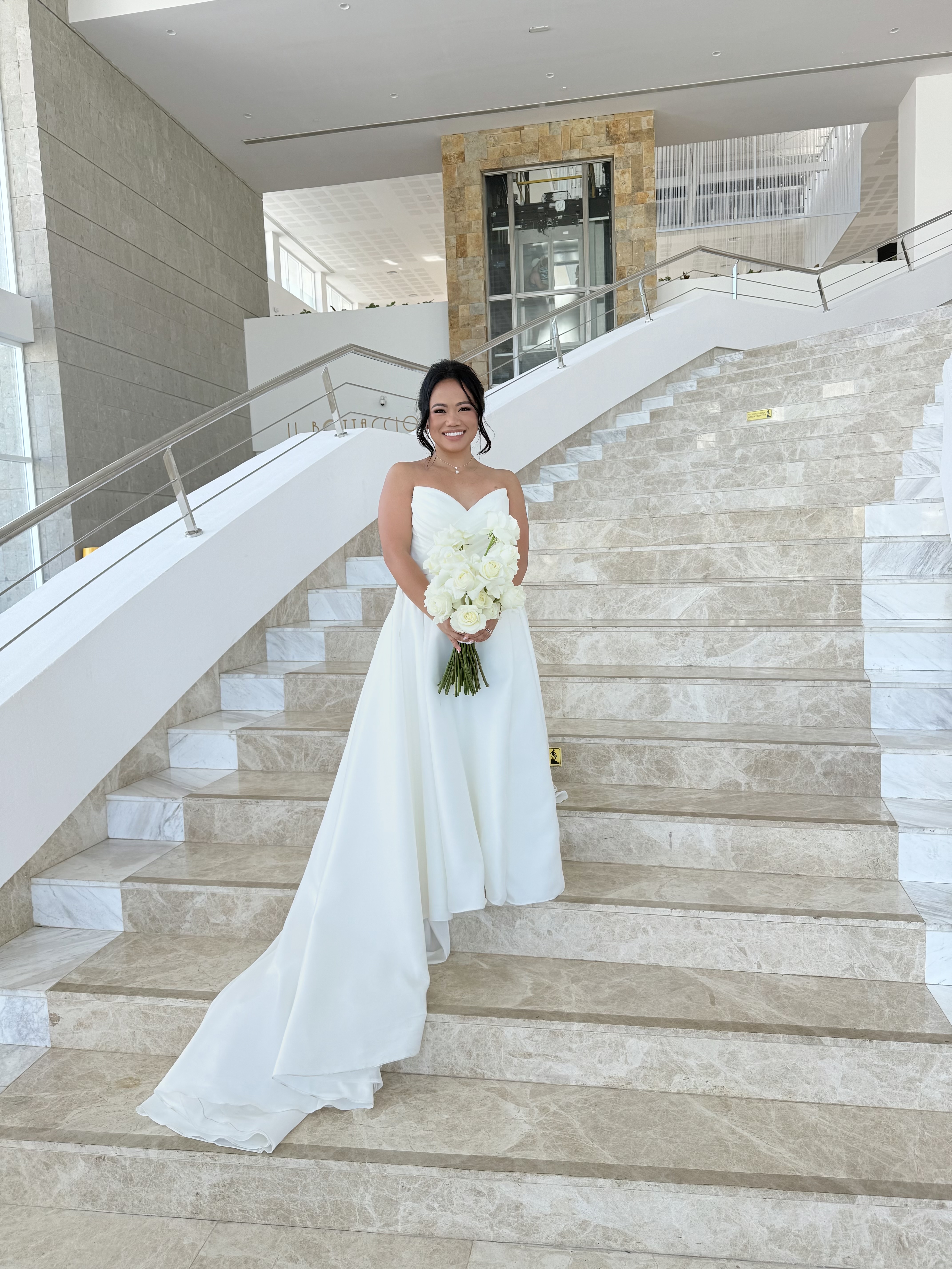 Bride in a white wedding dress holding a bouquet on elegant marble stairs, featuring Ashleigh and Dominic at their destination wedding.
