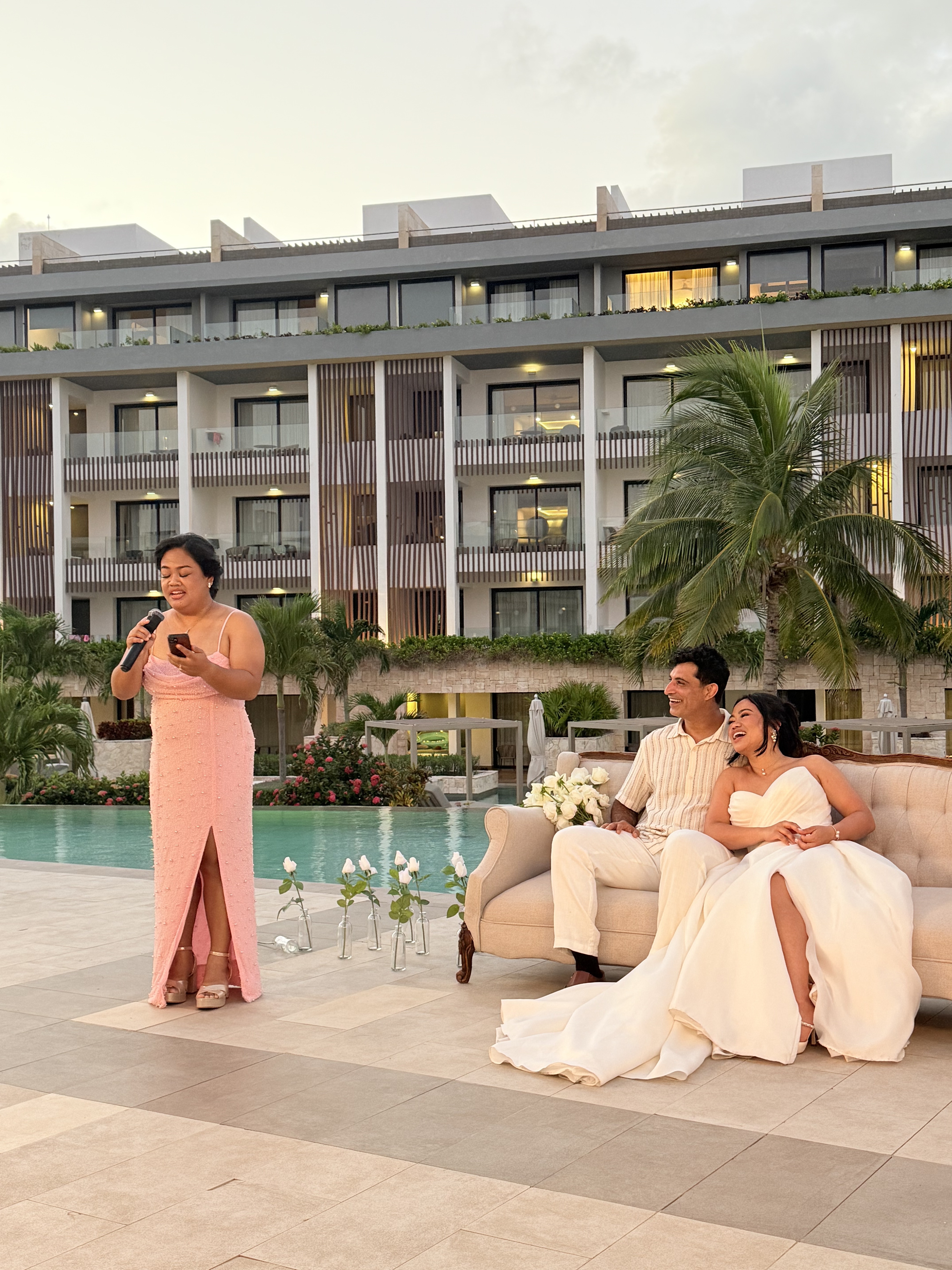 Ashleigh and Dominic beam in their wedding attire beside the pool, as a woman delivers a heartfelt speech during their destination wedding.