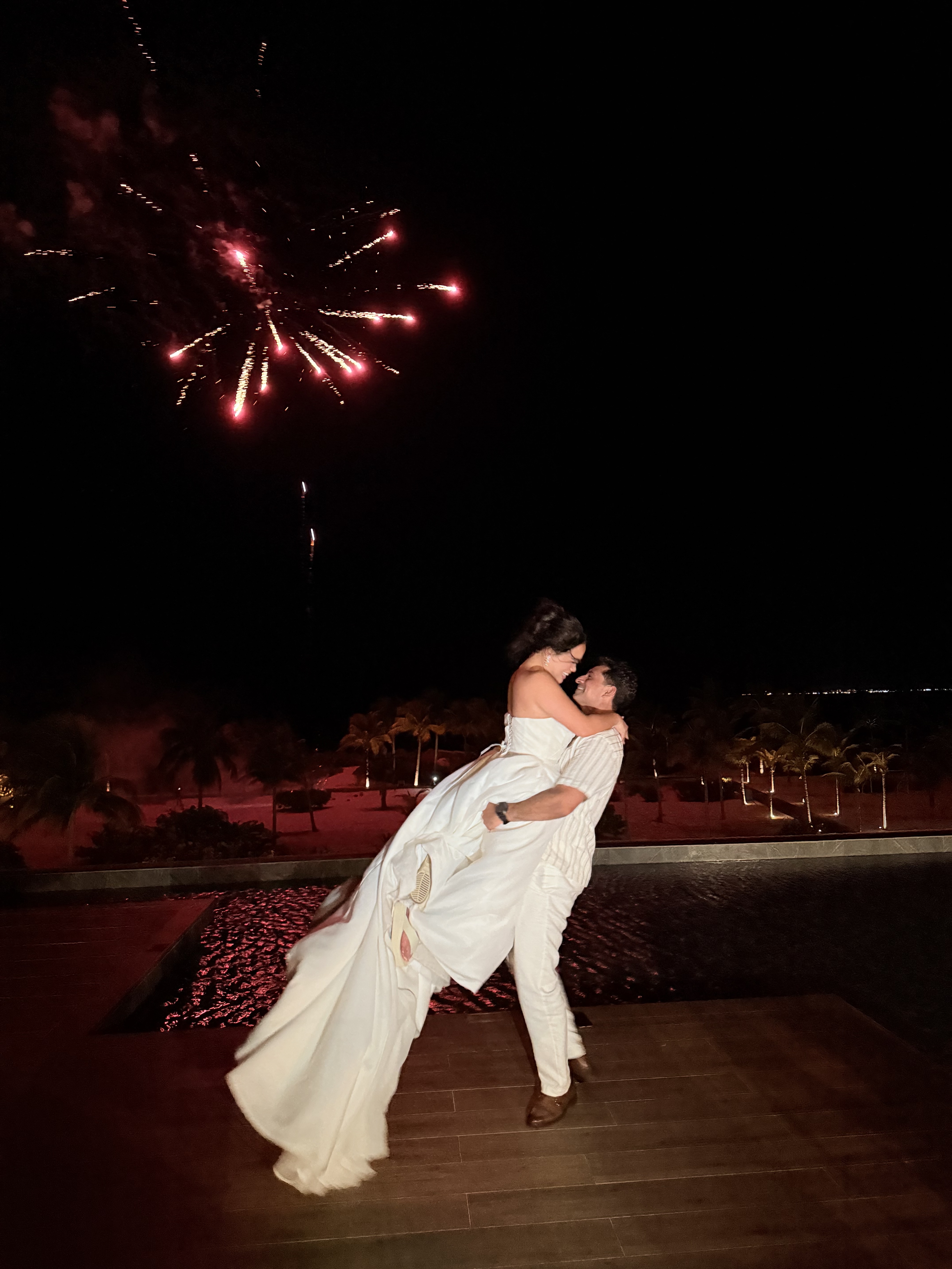 Ashleigh and Dominic, dressed in their wedding attire, embrace beneath a sky illuminated by fireworks during their unforgettable destination wedding.