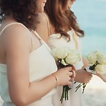 At a destination wedding, two women accessorize their white dresses with petite bouquets of roses at an outdoor celebration.