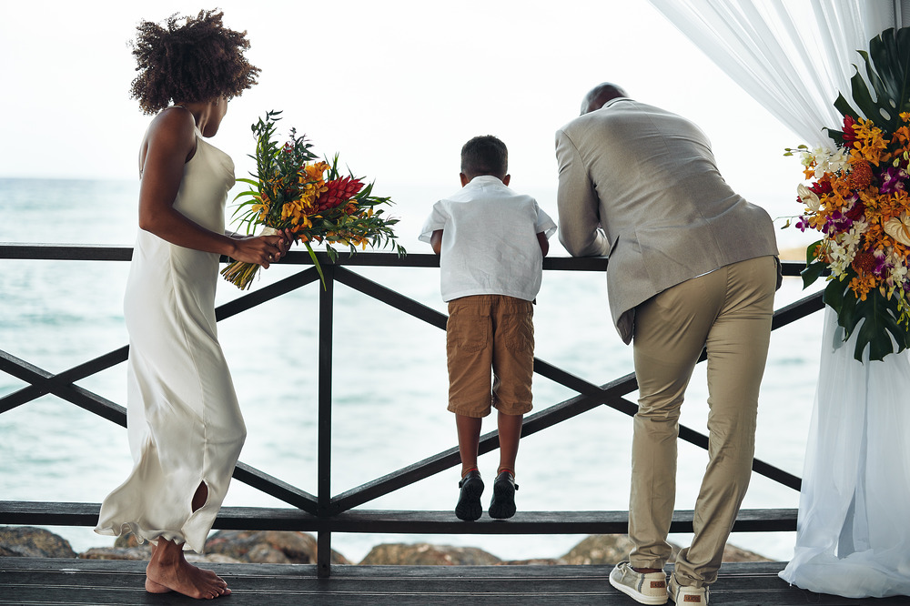 Three people stand by a wooden railing at a destination wedding, gazing out at the ocean with beautiful flower arrangements nearby.