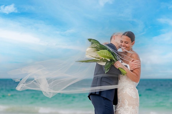 Bride and groom embrace on the beach during their destination wedding at Majestic Mirage, veil blowing.