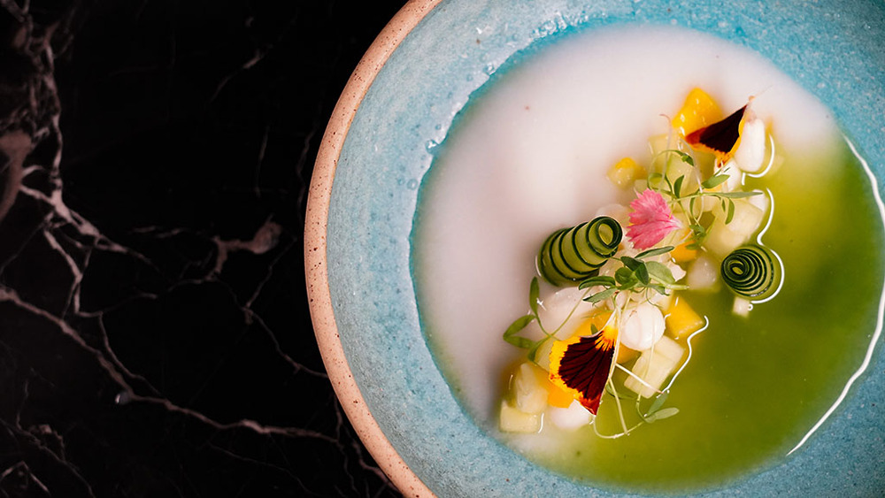 A blue ceramic bowl featuring an artistic arrangement of colorful vegetables and edible flowers in sauce, perfect for a destination wedding feast.