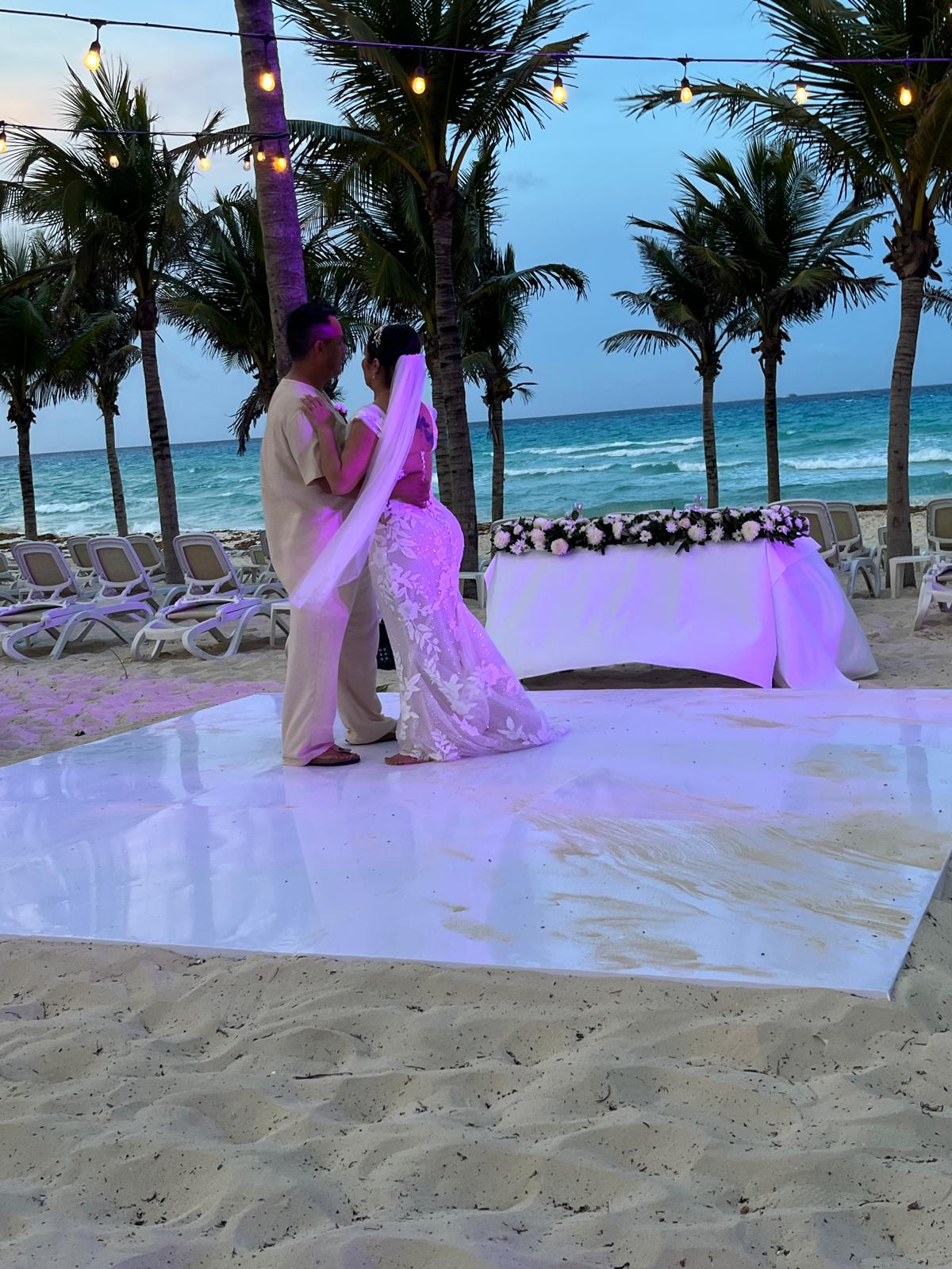 Camille and Jeff stand together on a beachside stage, palm trees swaying and the ocean stretching behind them, celebrating their beautiful destination wedding.
