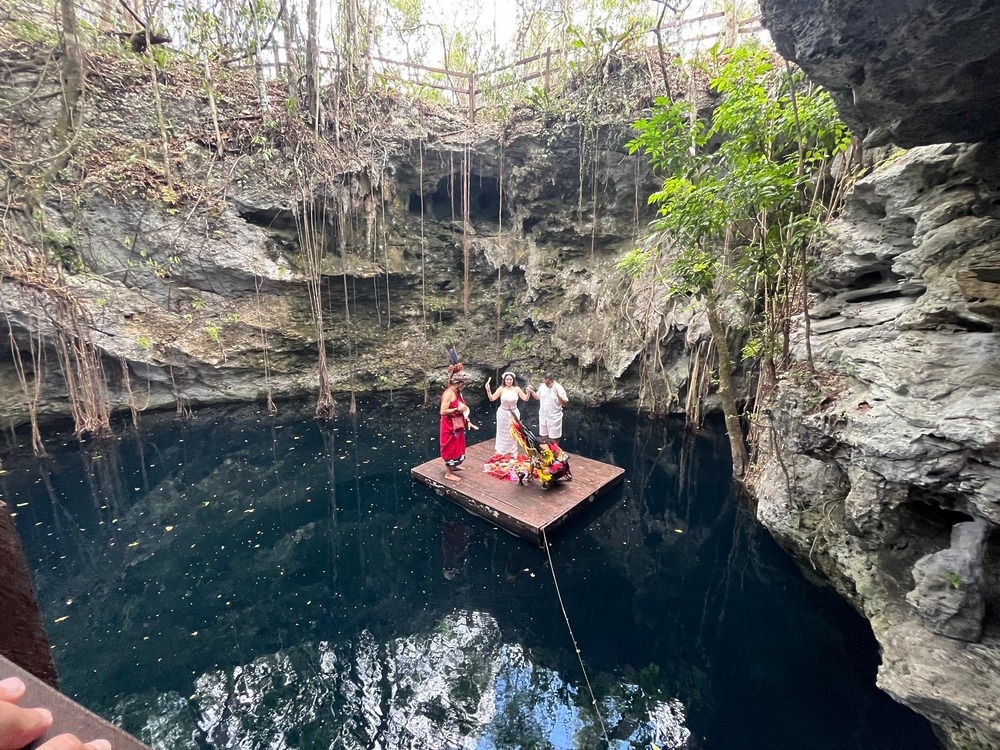Camille and Jeff, celebrating their destination wedding, stand on a wooden raft in a cenote surrounded by dramatic cliffs and lush vegetation.