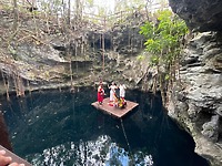 Camille and Jeff, celebrating their destination wedding, stand on a wooden raft in a cenote surrounded by dramatic cliffs and lush vegetation.
