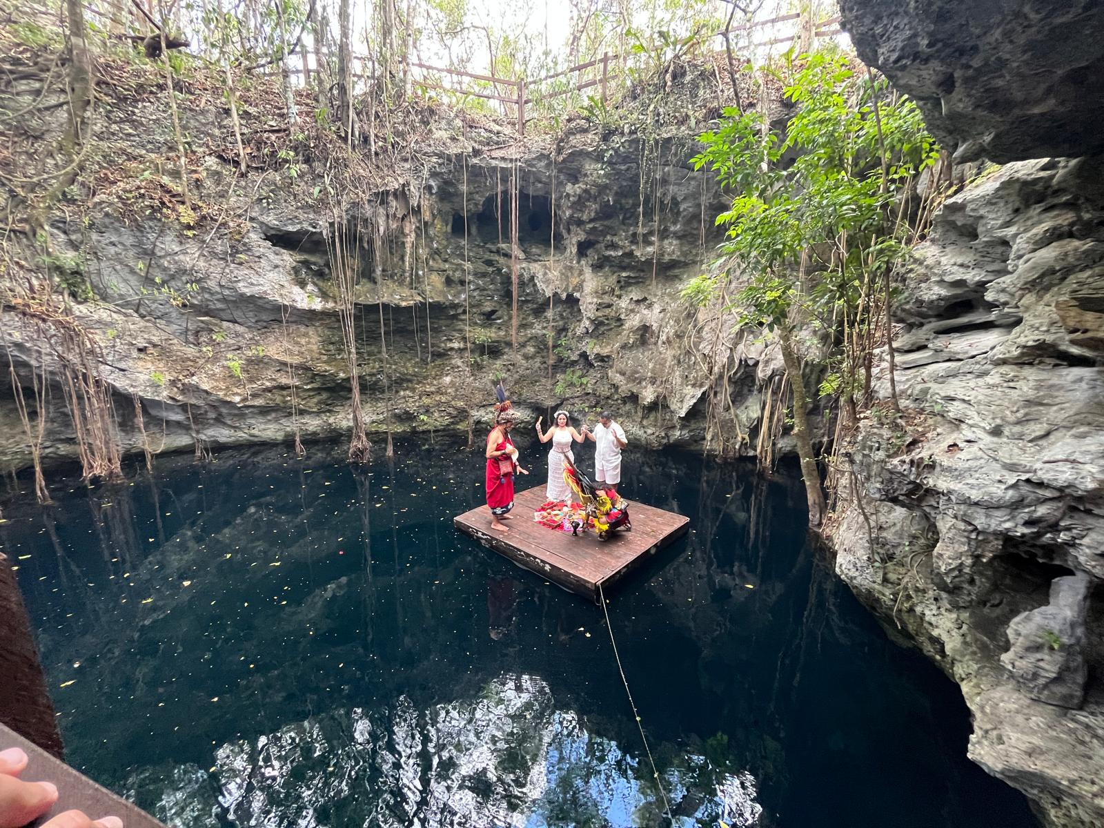 Camille and Jeff, celebrating their destination wedding, stand on a wooden raft in a cenote surrounded by dramatic cliffs and lush vegetation.