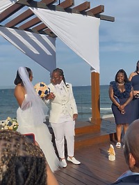 Natoya and Katrina hold hands at their destination wedding by the ocean, with guests looking on as they exchange vows.