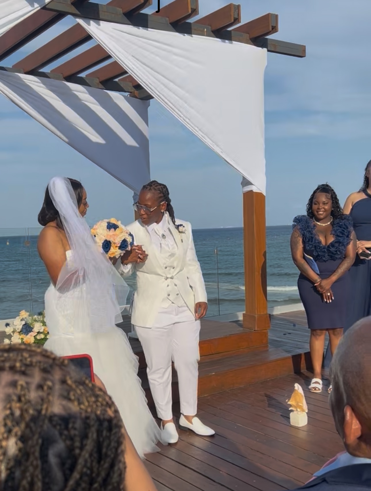 Natoya and Katrina hold hands at their destination wedding by the ocean, with guests looking on as they exchange vows.