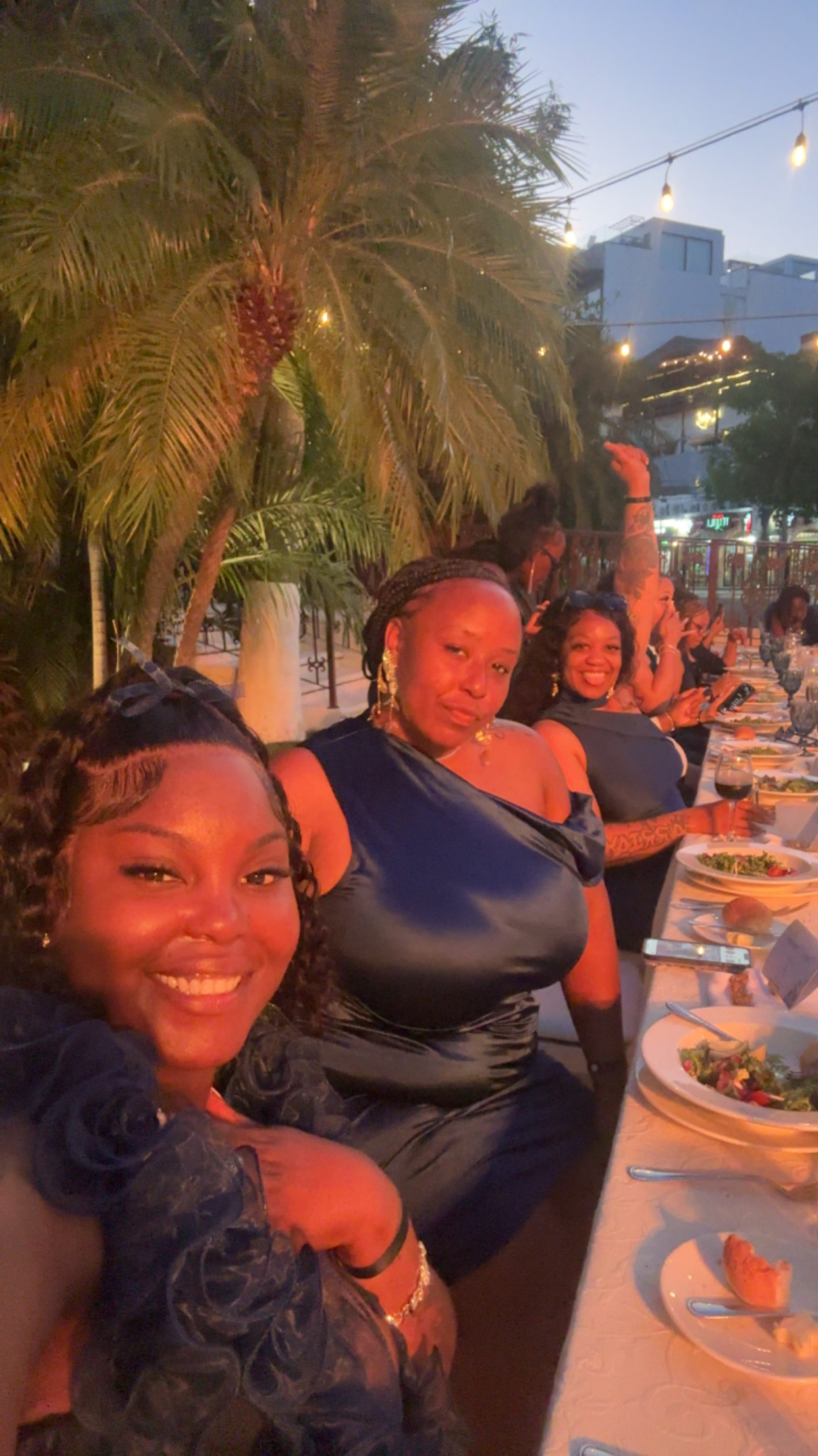 Women in elegant navy dresses, perfect for a destination wedding, dine outdoors with fresh salads, surrounded by palm trees.