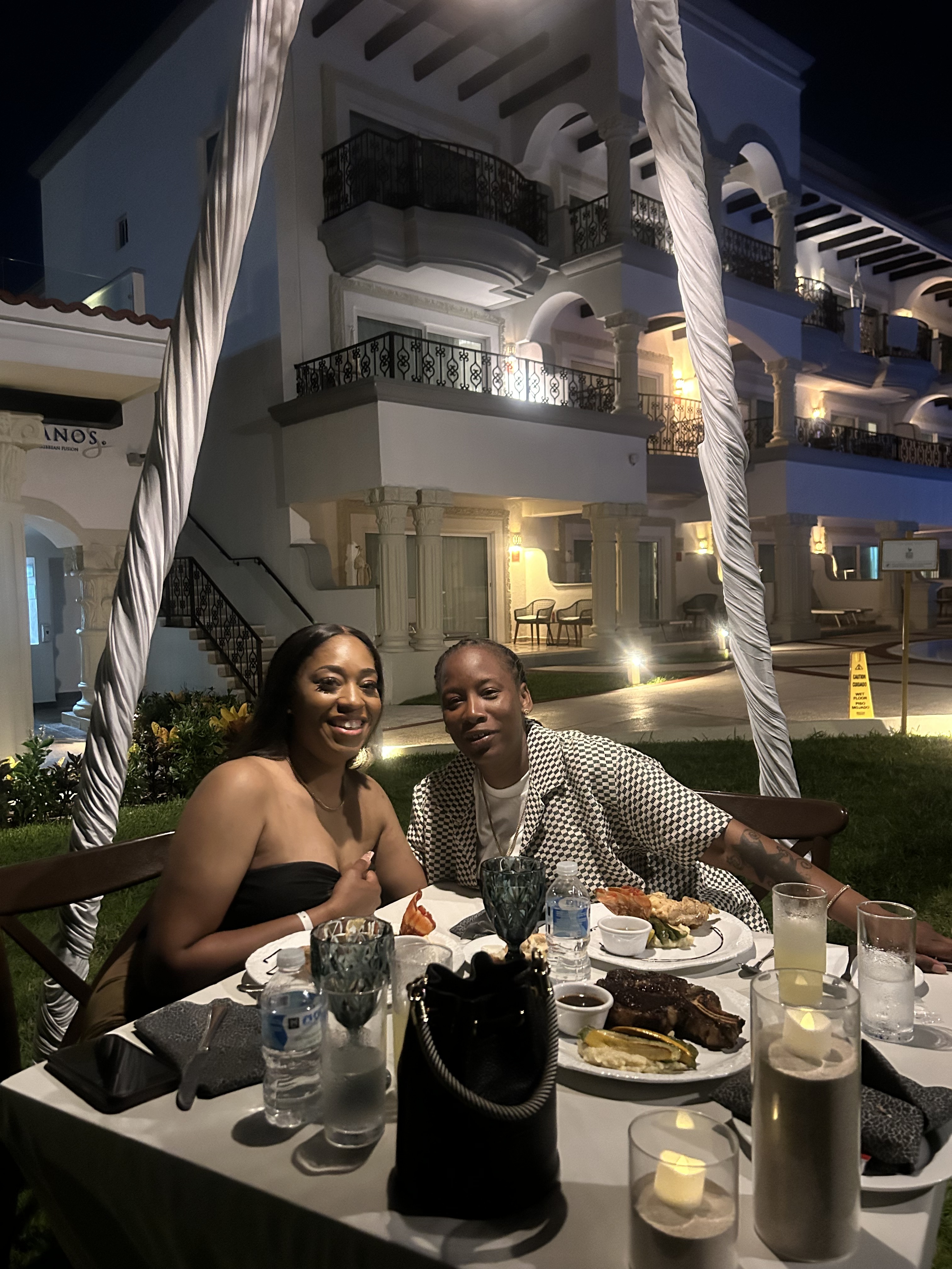 Grace and Mohammed sit at an outdoor dinner table at night, the glow of a multi-story building illuminating the backdrop of their destination wedding.