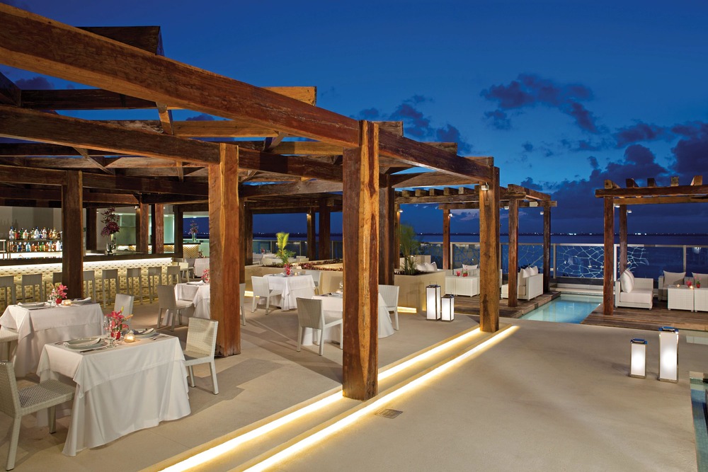 Seaside open-air restaurant at dusk, featuring wooden pergolas, white tables, and ambient lighting—an idyllic setting perfect for a destination wedding celebration.