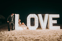 Aubrey and Noah pose beside a large illuminated "LOVE" sign on the sandy beach at night, capturing a magical moment from their destination wedding.