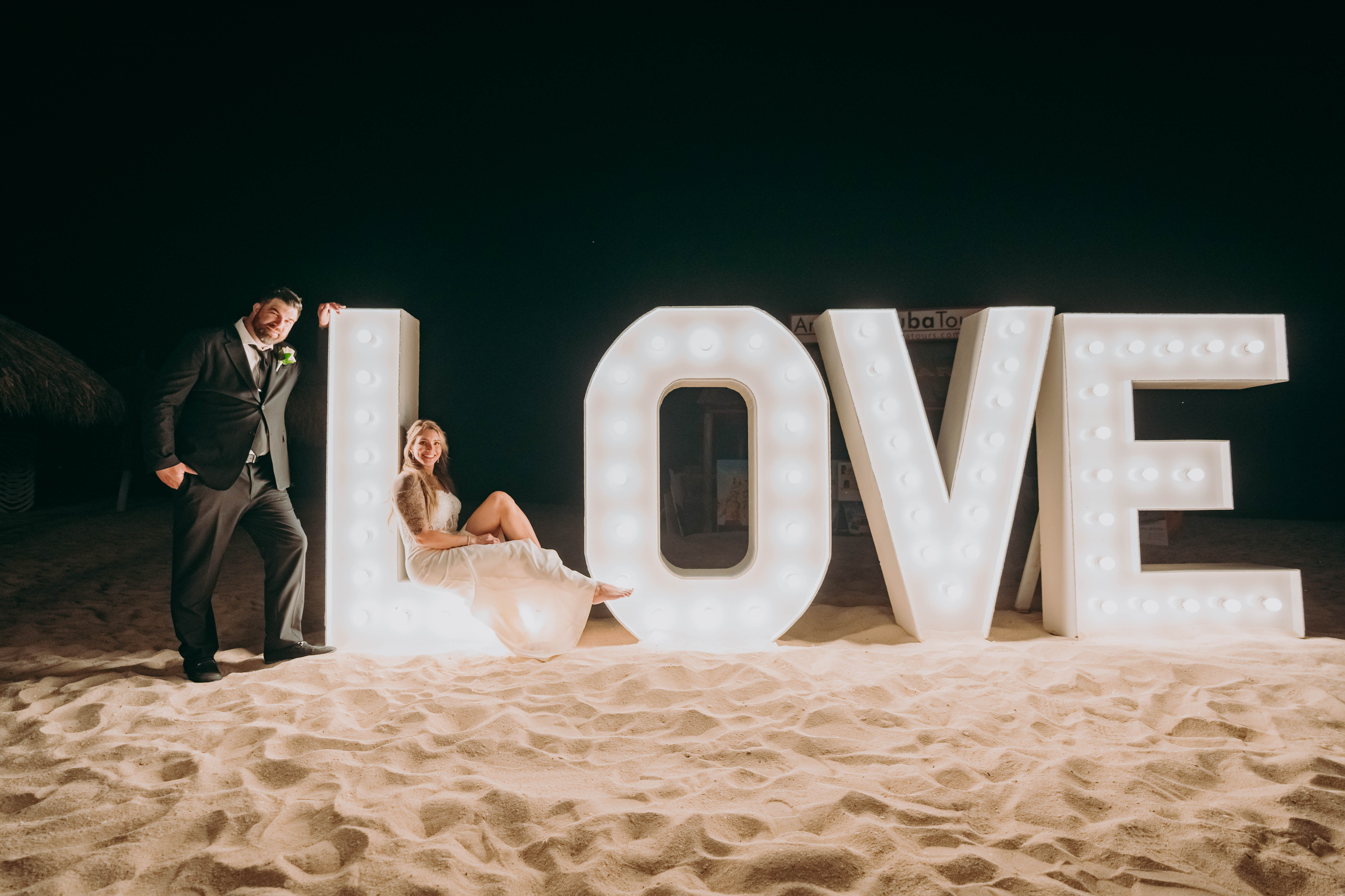 Aubrey and Noah pose beside a large illuminated "LOVE" sign on the sandy beach at night, capturing a magical moment from their destination wedding.