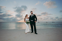 Aubrey and Noah stand side by side on a sandy beach at sunset, gazing into the distance as they celebrate their destination wedding.