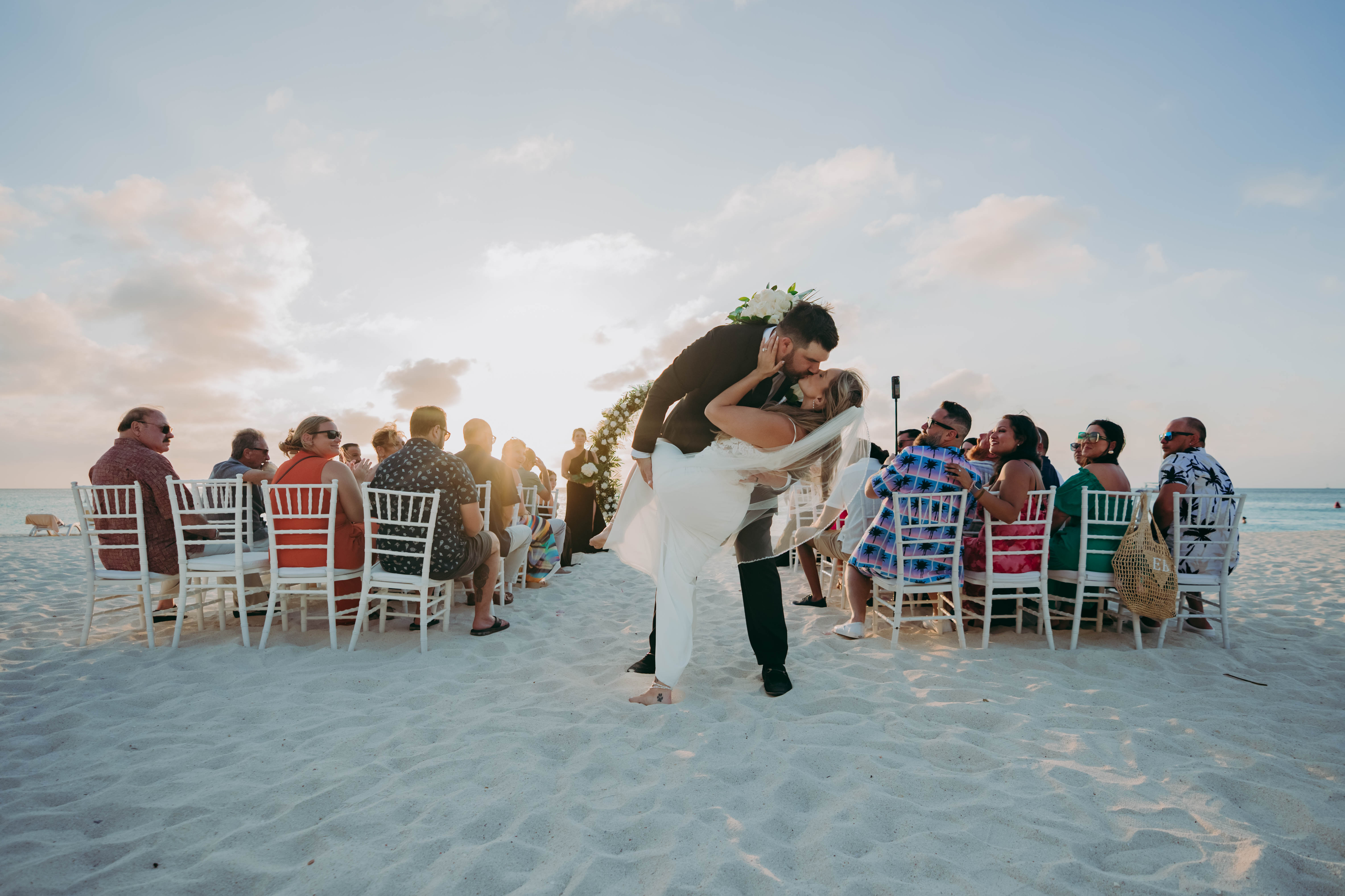 Aubrey and Noah share a kiss at their destination wedding on the beach, surrounded by loved ones as the sun sets.