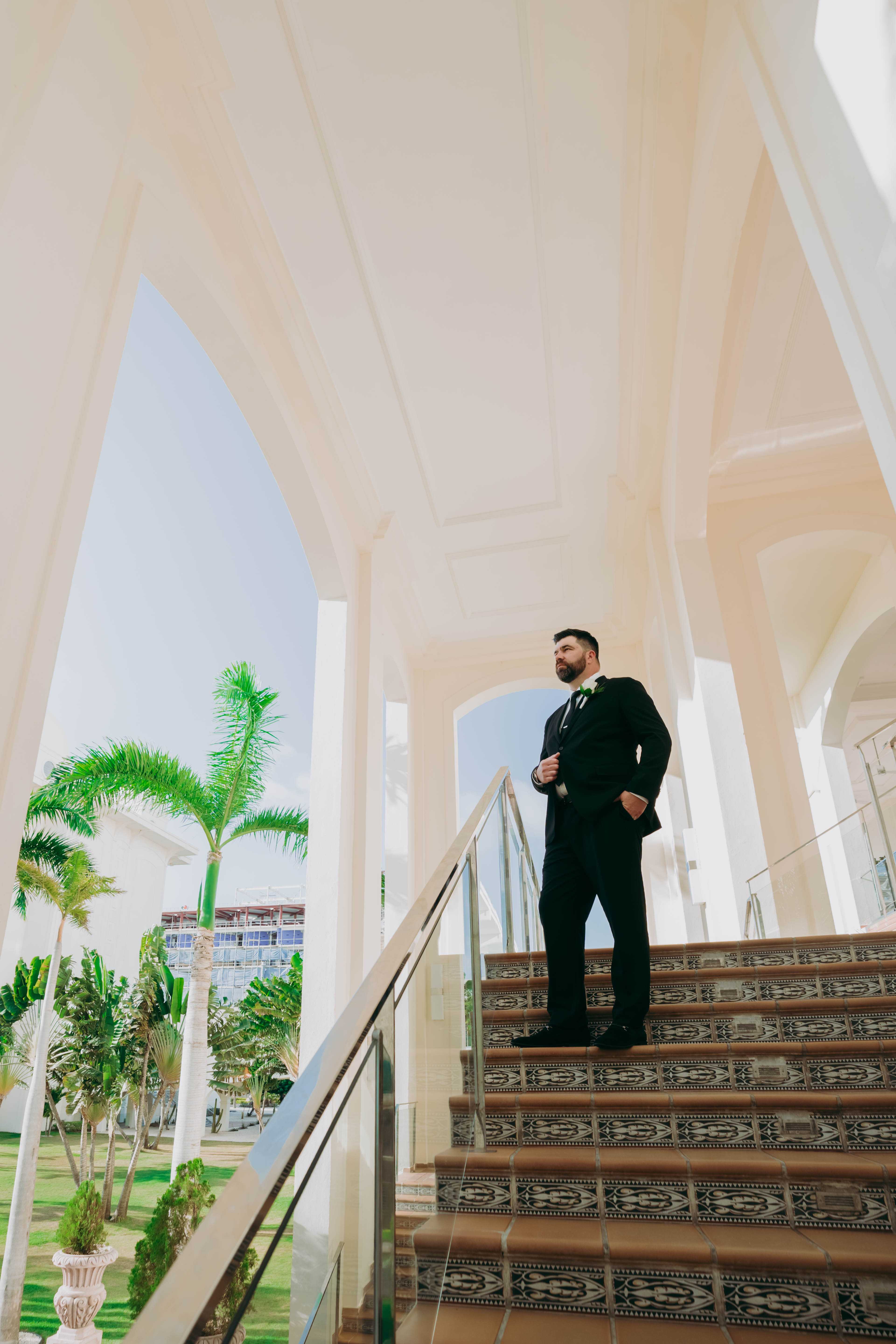 A man in a suit stands on tiled stairs beneath elegant white arches and swaying palm trees, capturing the essence of a destination wedding.