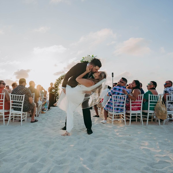 Aubrey and Noah share a kiss at the altar during their destination wedding on a sandy beach, surrounded by guests as the sun sets.