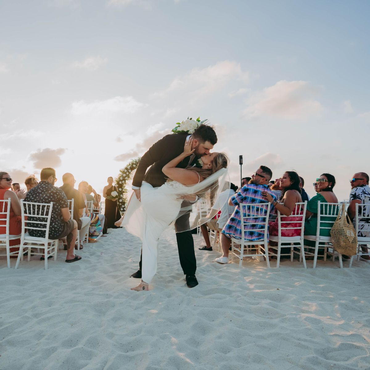 Aubrey and Noah share a kiss at the altar during their destination wedding on a sandy beach, surrounded by guests as the sun sets.