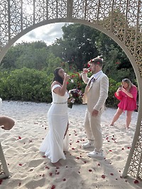 Aubrey and Noah, dressed in elegant white attire, raise a toast on a sandy beach adorned with scattered petals, celebrating their destination wedding.